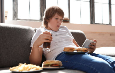 Teen boy eating junk food, drinking soda while looking at smartphone