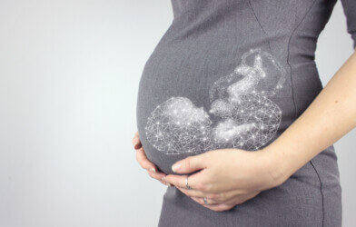 Pregnant woman in dress holds hands on belly with hologram fetus on a grey background.